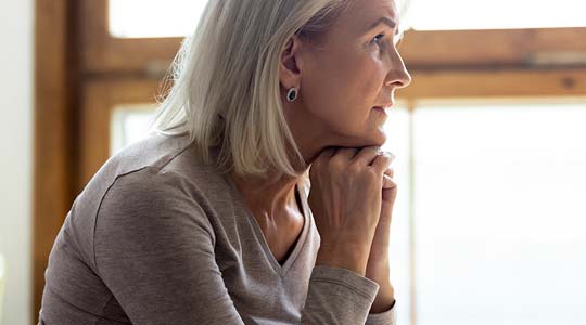 Trist, midaldrende kvinde sidder med hænderne under hagen og kigger ud af vinduet.