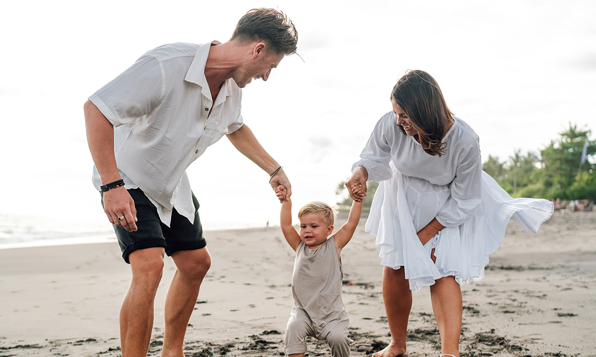 En far og mor holder et lille barn i hånden på en strand.