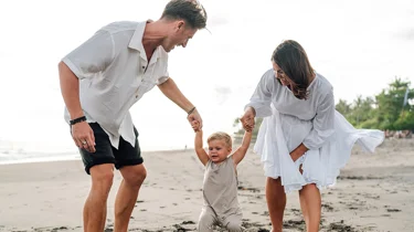 En far og mor holder et lille barn i hånden på en strand.