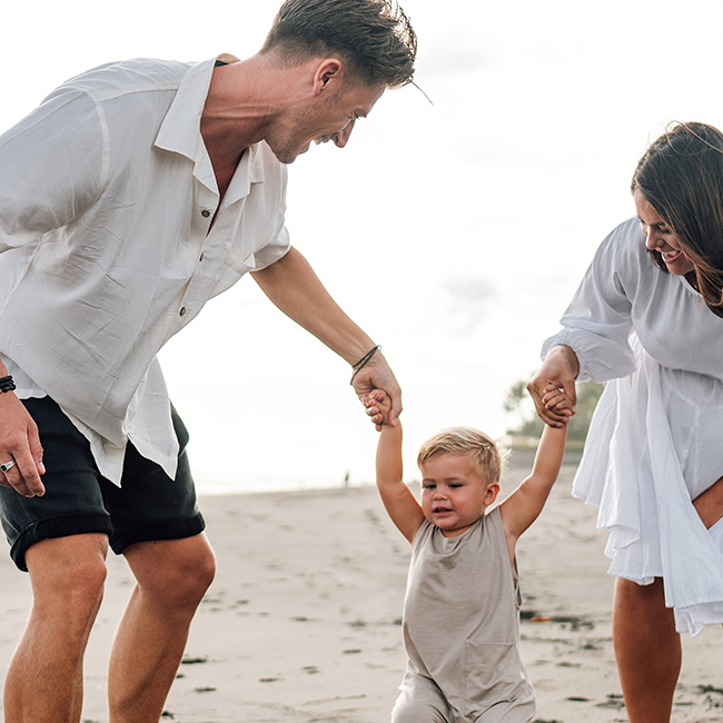 En far og mor holder et lille barn i hånden på en strand.