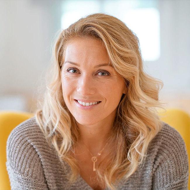 Kvinde 40+ sidder i en gul stol og smiler in i kameraet.