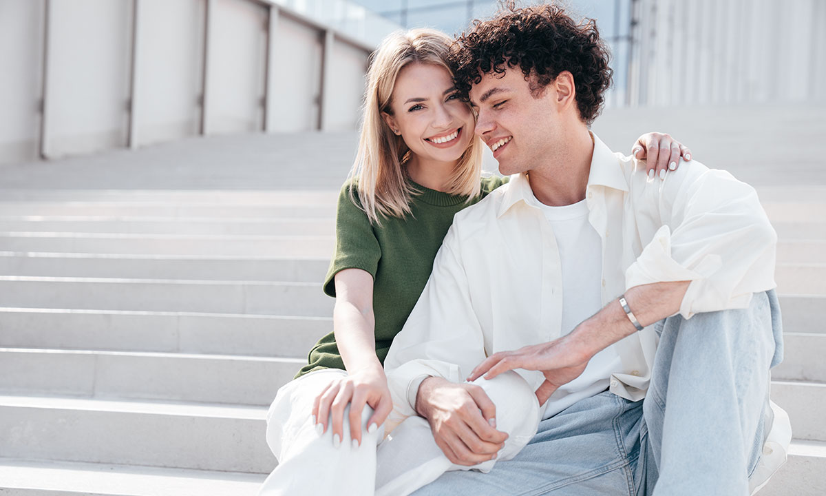 Et ungt par holder on hinanden på en trappe - hun smiler ind i kameraet mens han ser ned.