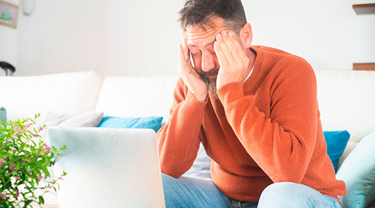 Træt mand sidder i en sofa med fingrene i tindingerne og ser ned på sin computer.