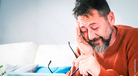 Træt mand sidder i en sofa med fingrene i tindingerne og ser ned på sin computer.