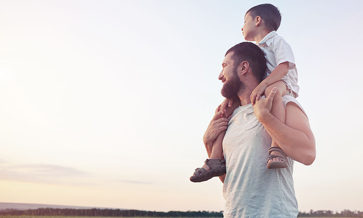 Far står med sin søn på skuldrene ude i naturen.