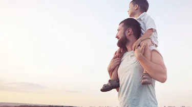 Far står med sin søn på skuldrene ude i naturen.