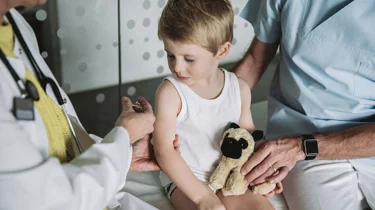 Et barn er ved at få en vaccination i trygge omgivelser og med sin bamse på skødet.
